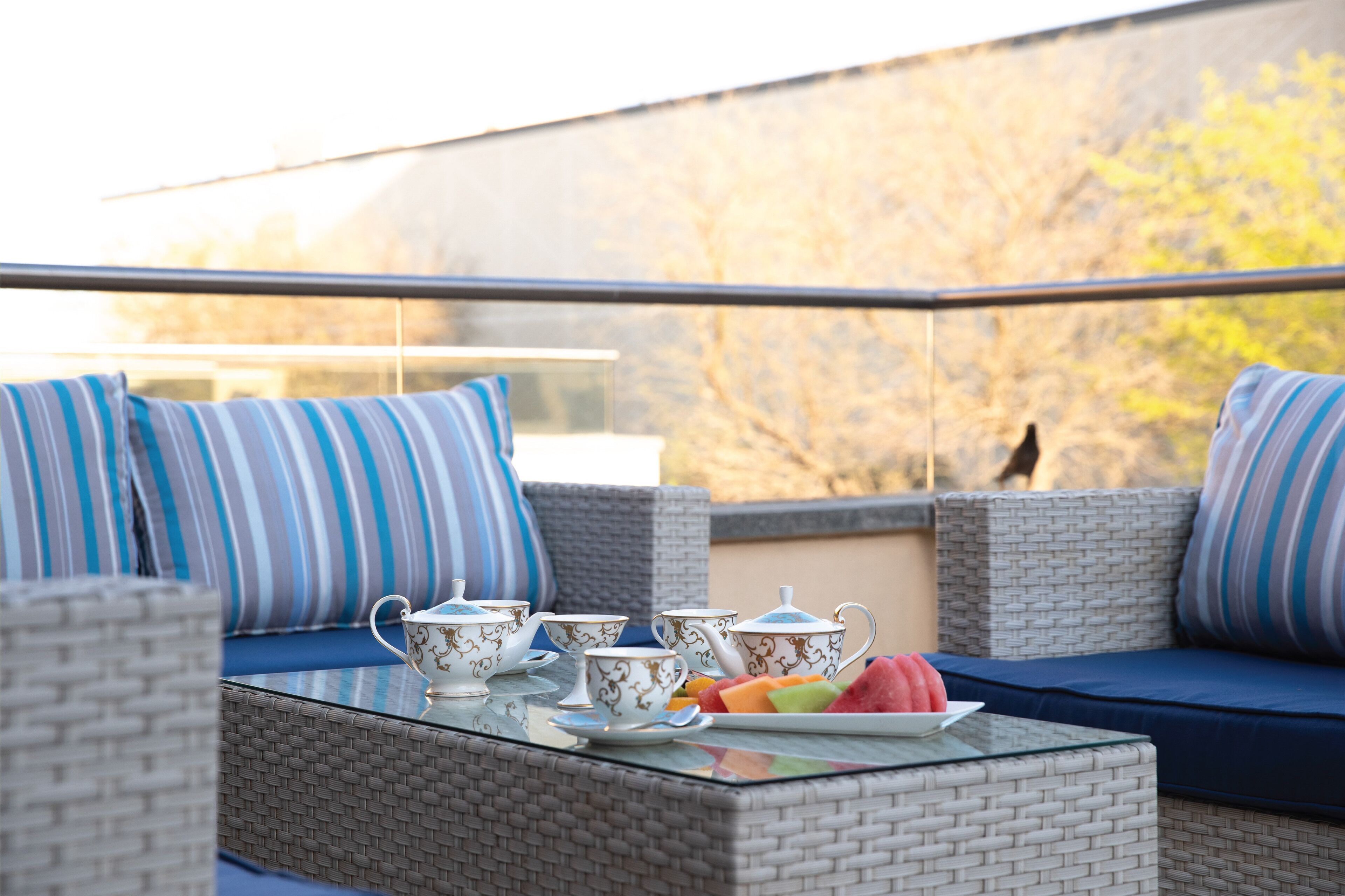 A balcony with wicker furniture, striped cushions, and a table with teapots and fruit.