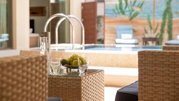 A poolside area with wicker furniture, a glass table with fruits and glasses, and a view of the pool and beach through a large window.