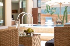 Un espace aménagé au bord de la piscine, avec des meubles en osier, une table en verre sur laquelle sont disposés des fruits et des verres, et une vue sur la piscine et la plage, visible à travers une grande fenêtre.