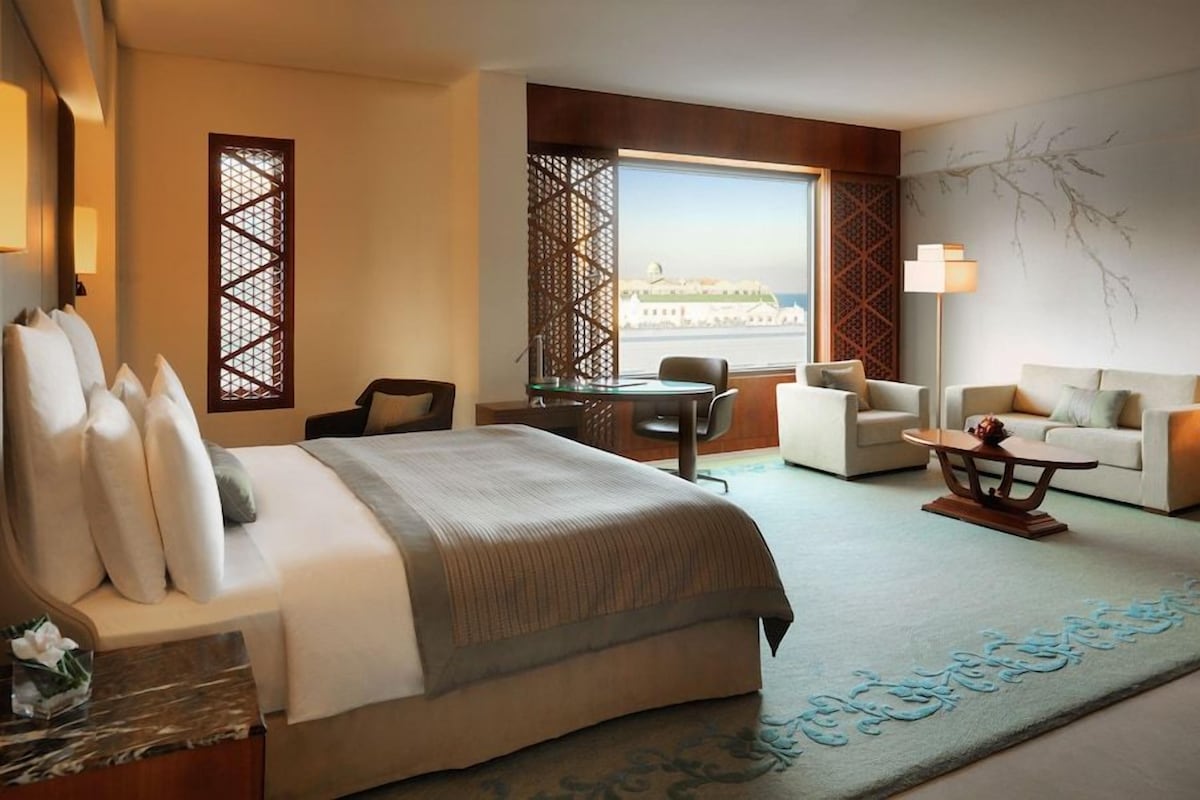 A hotel room with a bed, two armchairs, a sofa, a small table, and a desk. There is a window with a view of a building and a tree.