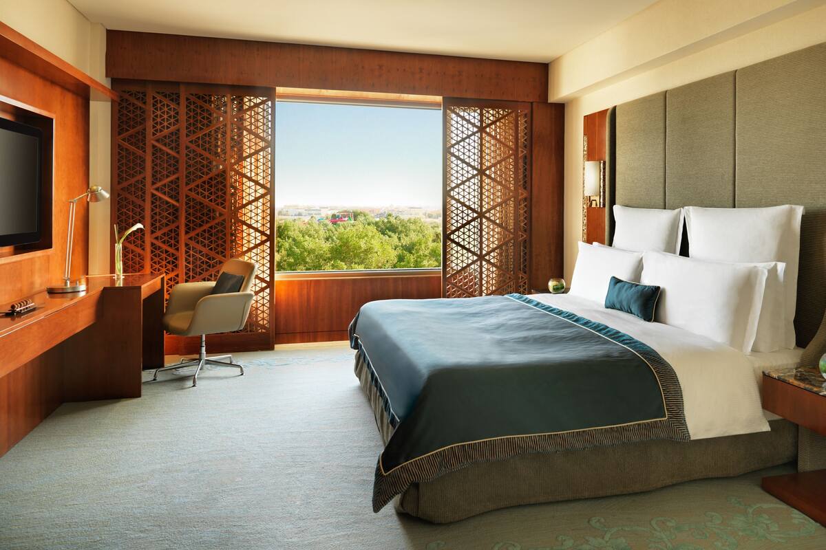 A hotel room with a large bed, a desk with a chair, a television, and a view of greenery through a window.