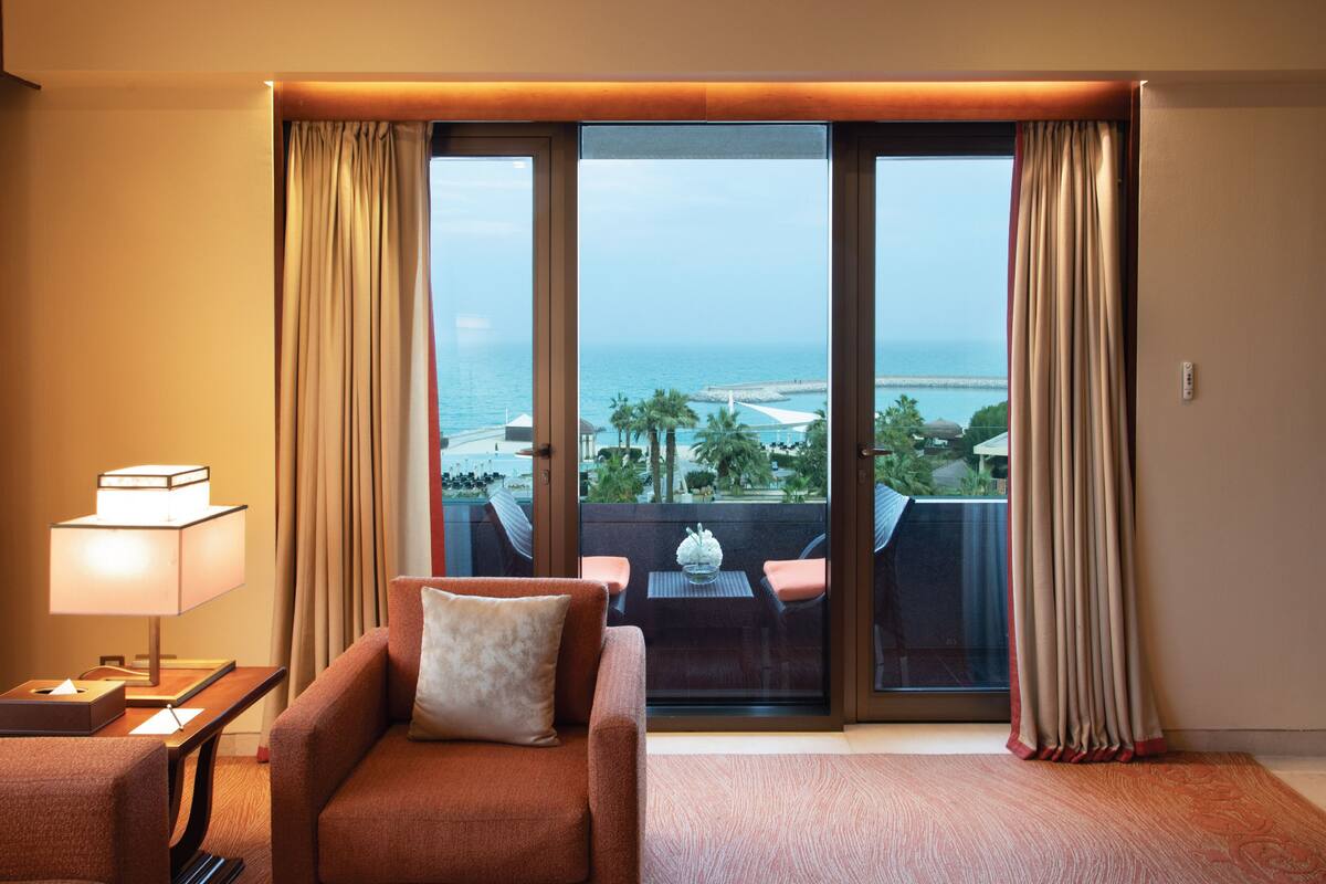 A hotel room with a sofa, a table with a lamp, and a view of the sea through large glass doors.