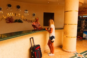A reception desk with a sign reading "HILLS RESORT" and a clock above it. A suitcase is placed on the floor beside the desk.