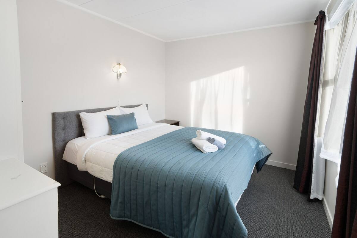 A hotel room with a bed, a bedside table, a lamp, and a window with curtains.