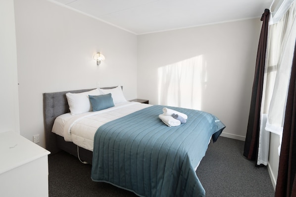 A hotel room with a bed, a bedside table, a lamp, and a window with curtains. - Oakleigh Motel (Gore)