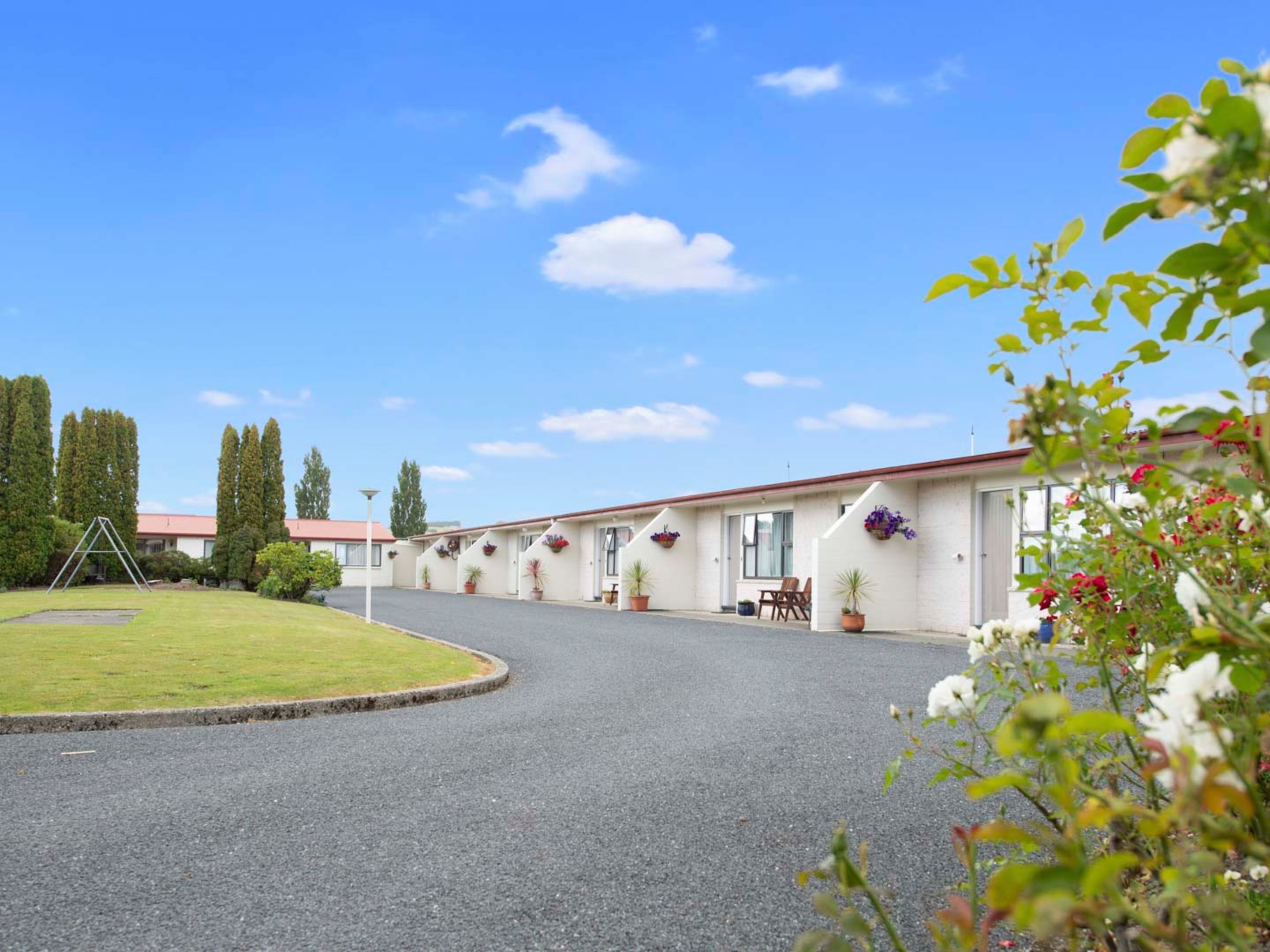 A row of single-story mobile homes with individual patios, surrounded by a well-maintained lawn and tall trees.