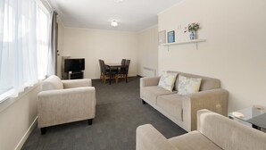 Uma sala de estar com sofá, poltronas, televisão, mesa de jantar com cadeiras e uma pequena estante com objetos decorativos.