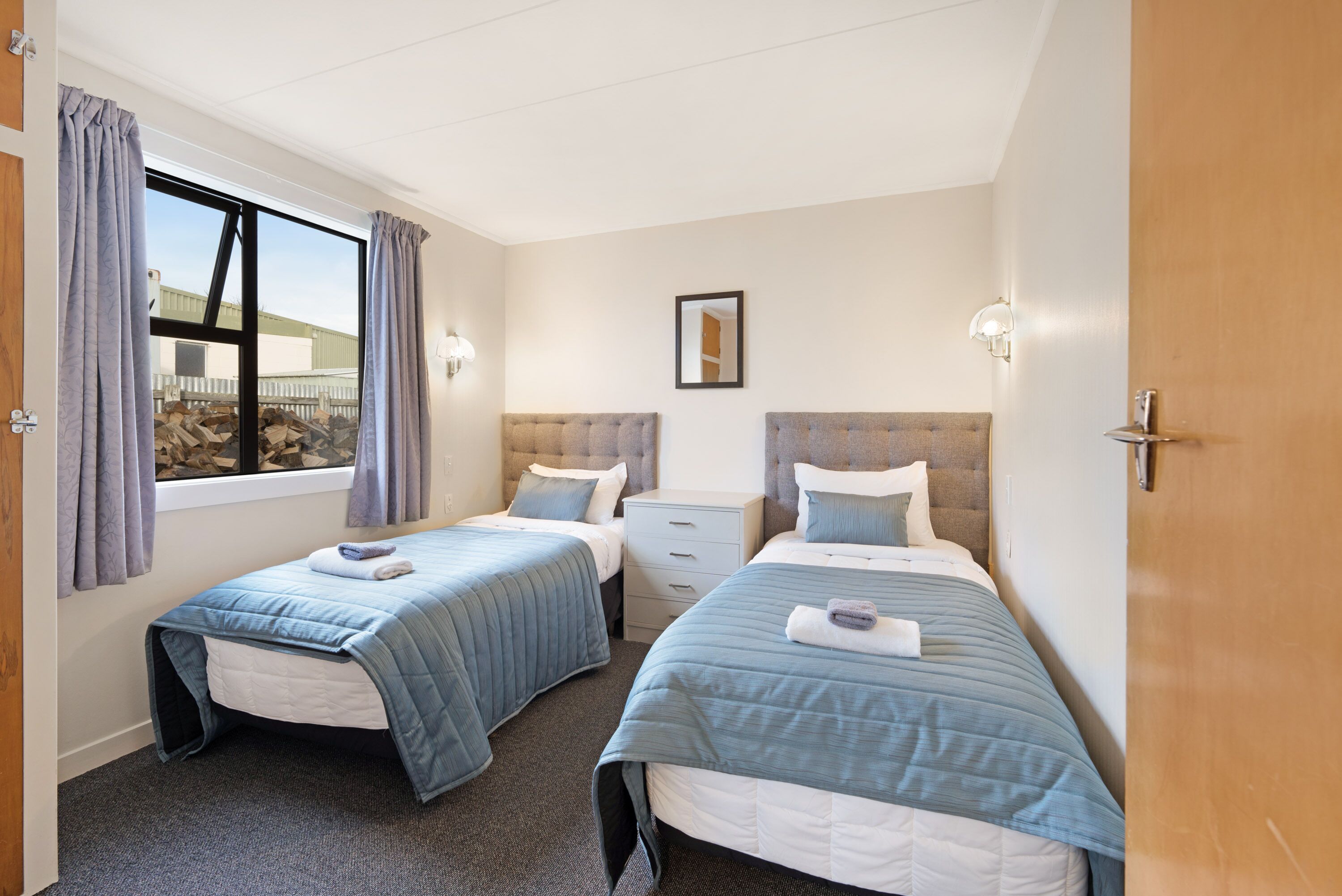 A hotel room with two single beds, a window with curtains, a bedside table with a lamp, and a view of a building outside.