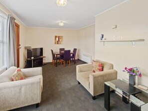 A living room with two sofas, a TV, a dining table with chairs, a small table with a vase of flowers, and a ceiling fan. - Oakleigh Motel (Gore)