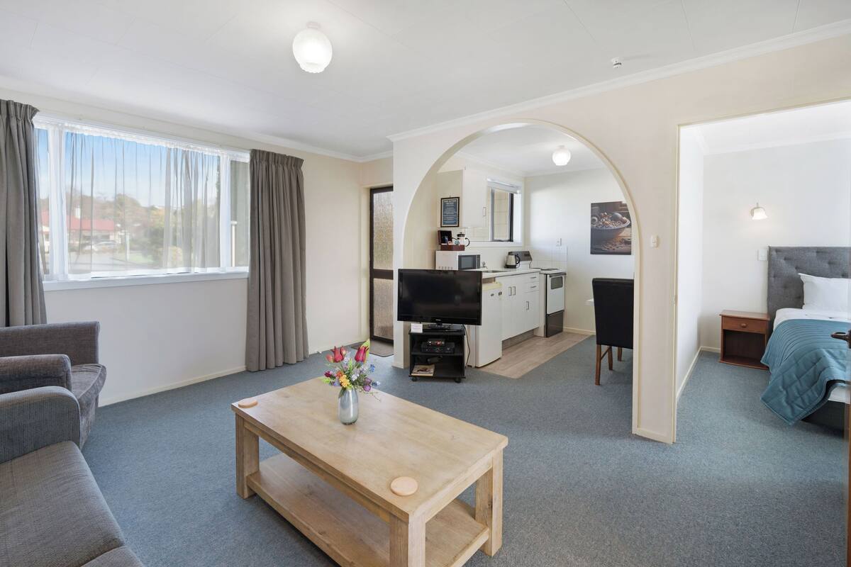 A modern hotel room with a sofa, a wooden table with a vase of flowers, a TV, a kitchenette, and a bed.