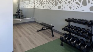 Un gimnasio con suelo de madera, una banca, un espejo adosado a la pared y un estante con mancuernas.
