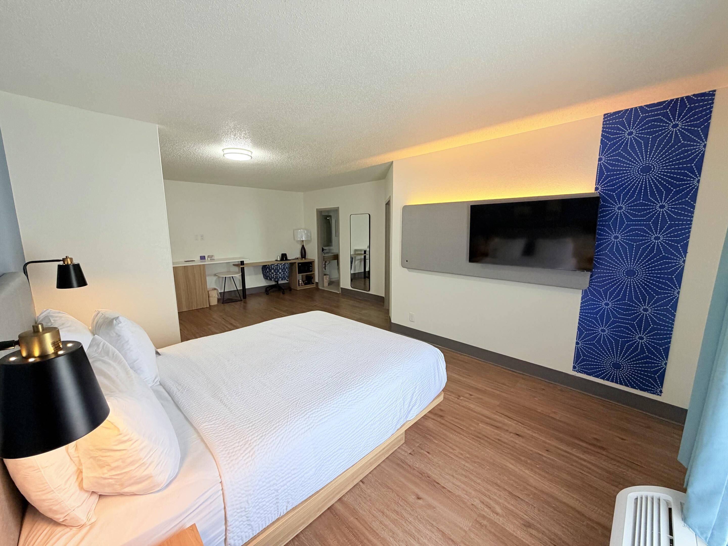 A hotel room with a bed, a desk, a mirror, a TV, and a patterned wall.