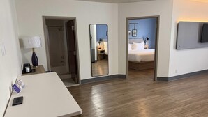 A hotel room with a wooden floor, a desk with a lamp, a bed, a mirror, and a TV mounted on the wall.