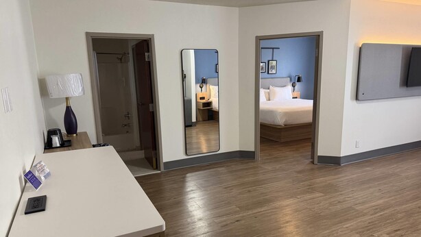 Habitación de hotel con suelo de madera, un escritorio con lámpara, cama, espejo y televisor montado en la pared.