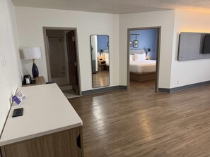 A hotel room with a wooden floor, a desk with a lamp, a bed, a mirror, and a TV mounted on the wall.