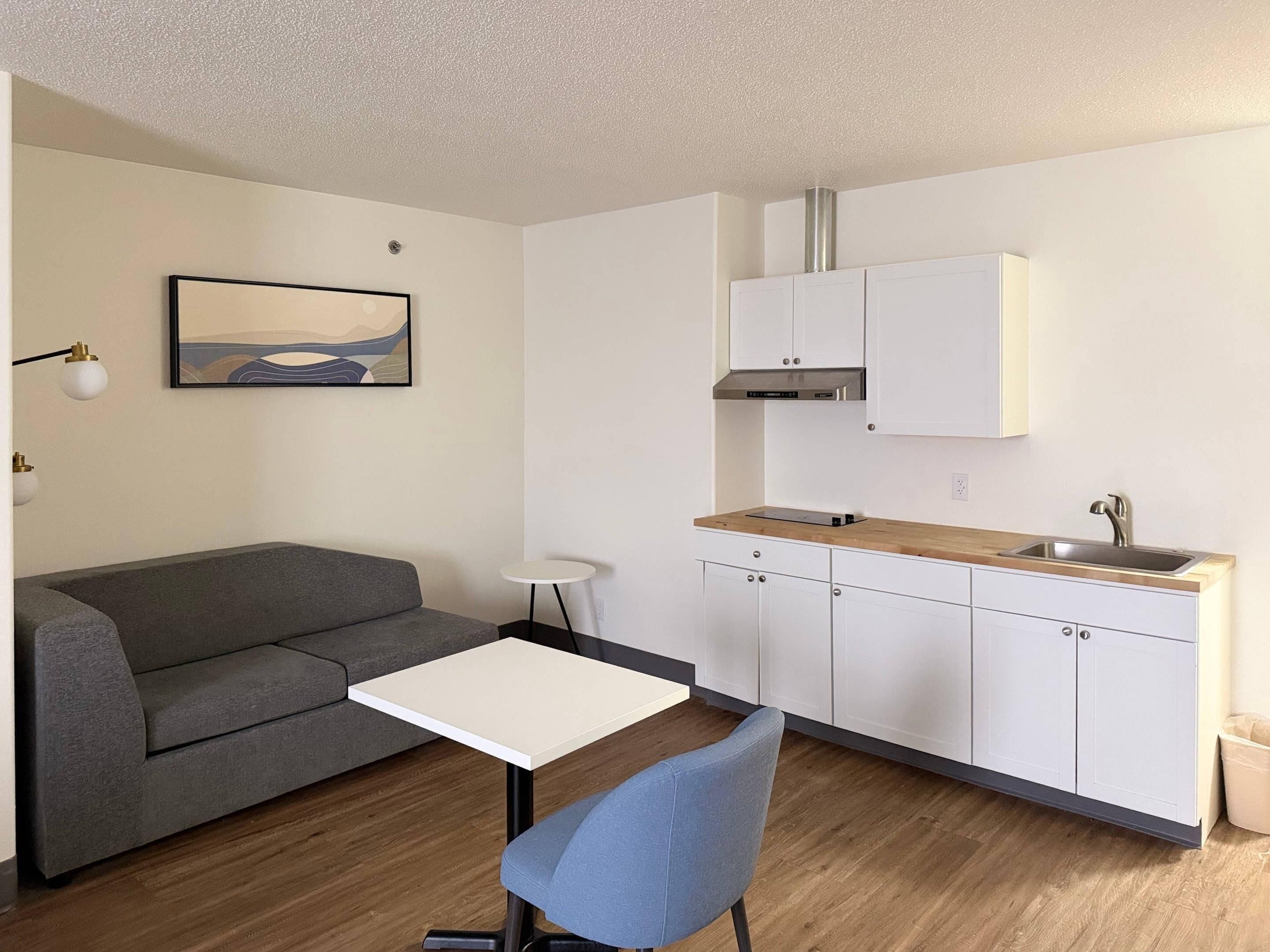 A compact living space with a grey sofa, a small white table, a blue chair, a kitchenette with white cabinets, and a painting on the wall.