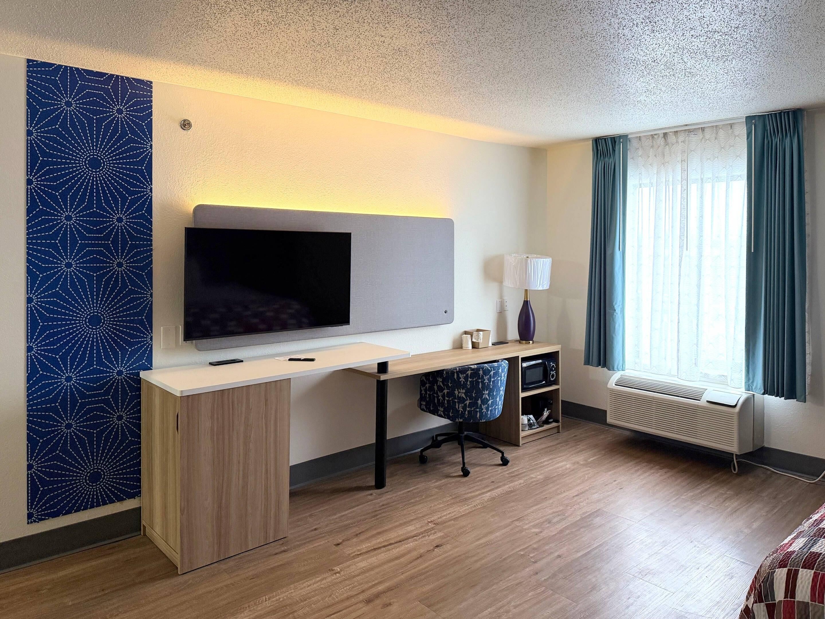 A hotel room with a flat-screen TV, a desk, a chair, a lamp, a heater, and a window with curtains.