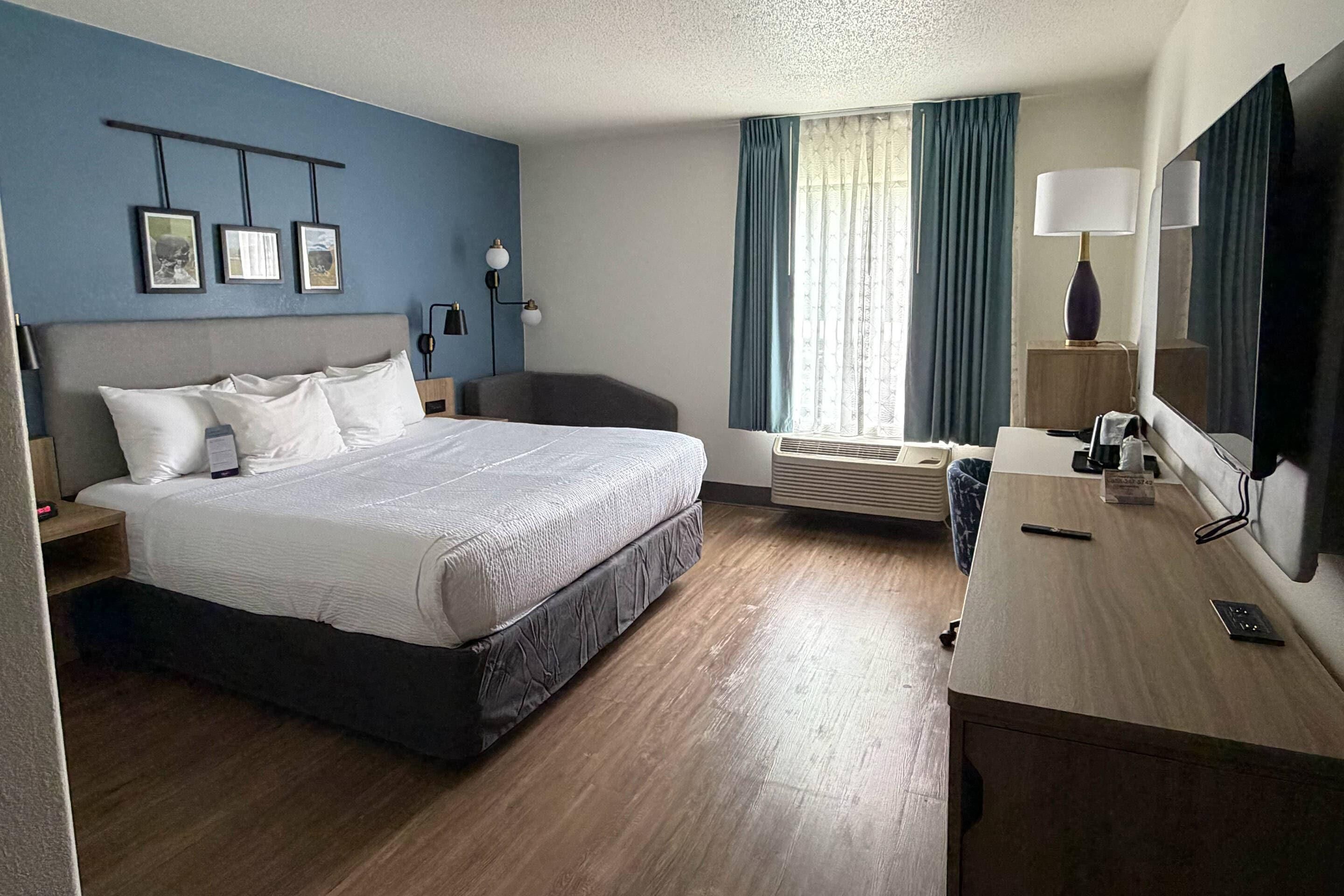 A hotel room with a bed, desk, chair, air conditioning unit, and a TV.