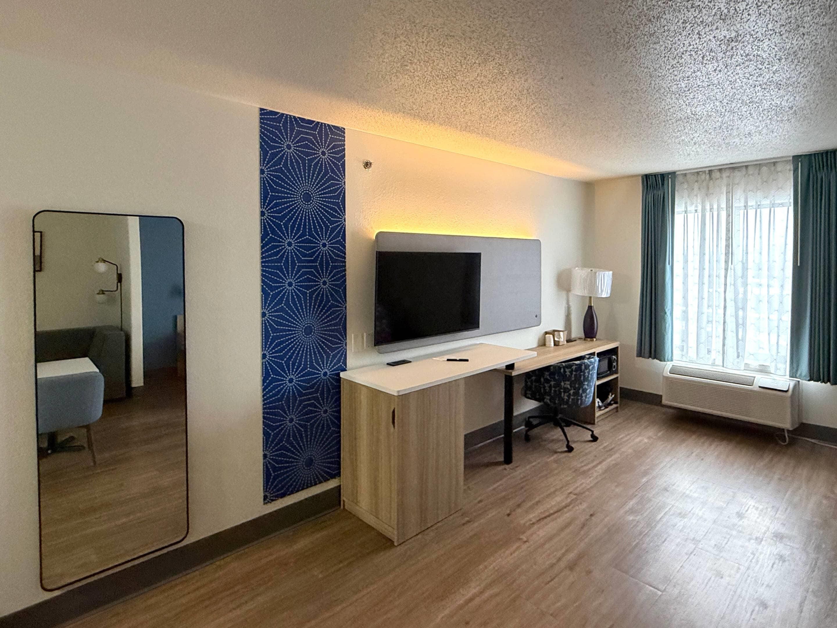 Habitación de hotel con un espejo grande, televisor de pantalla plana, escritorio con lámpara, una silla y ventana con cortinas.