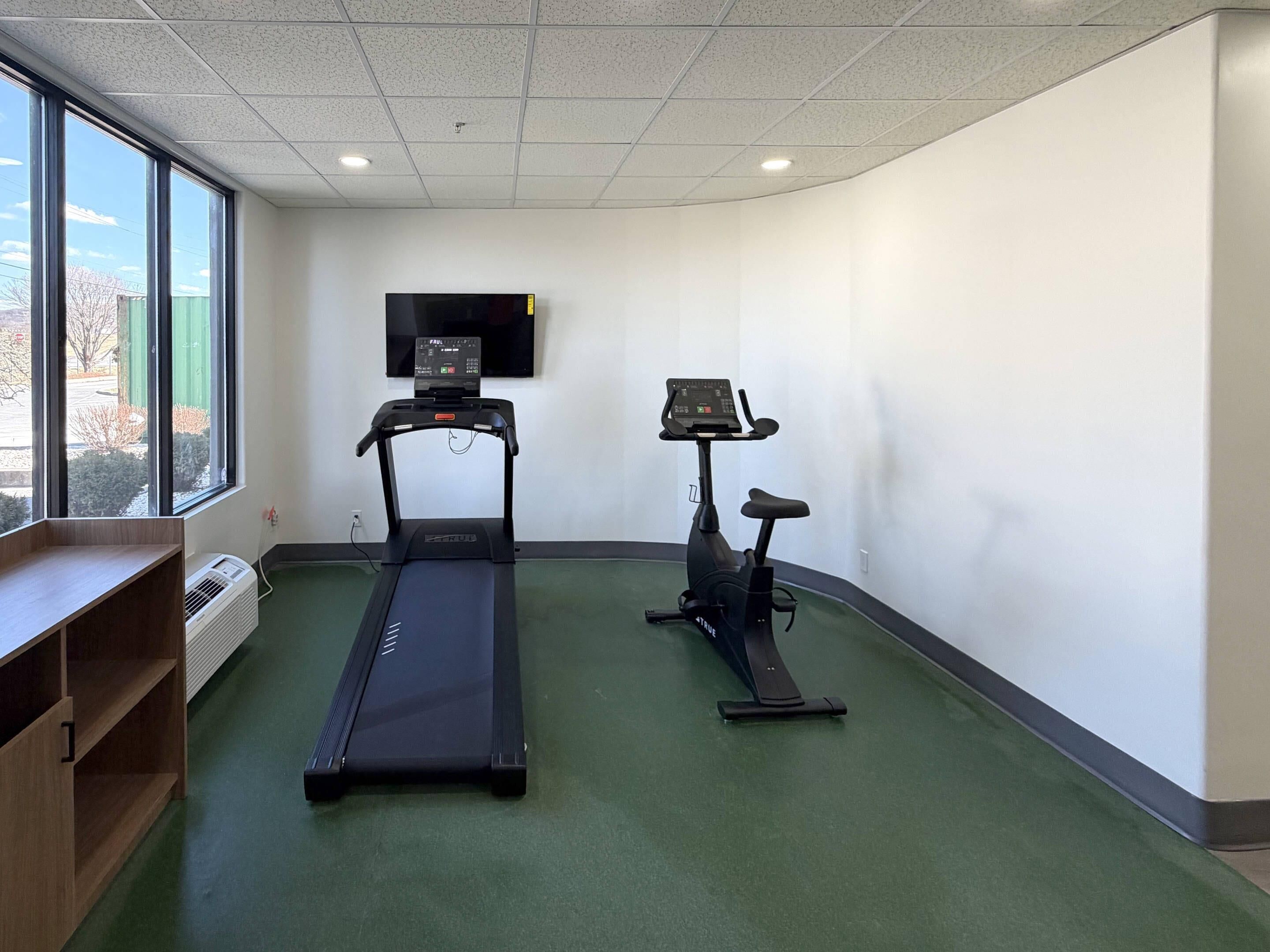 A fitness room with a treadmill and an elliptical machine, a TV on the wall, and a large window.