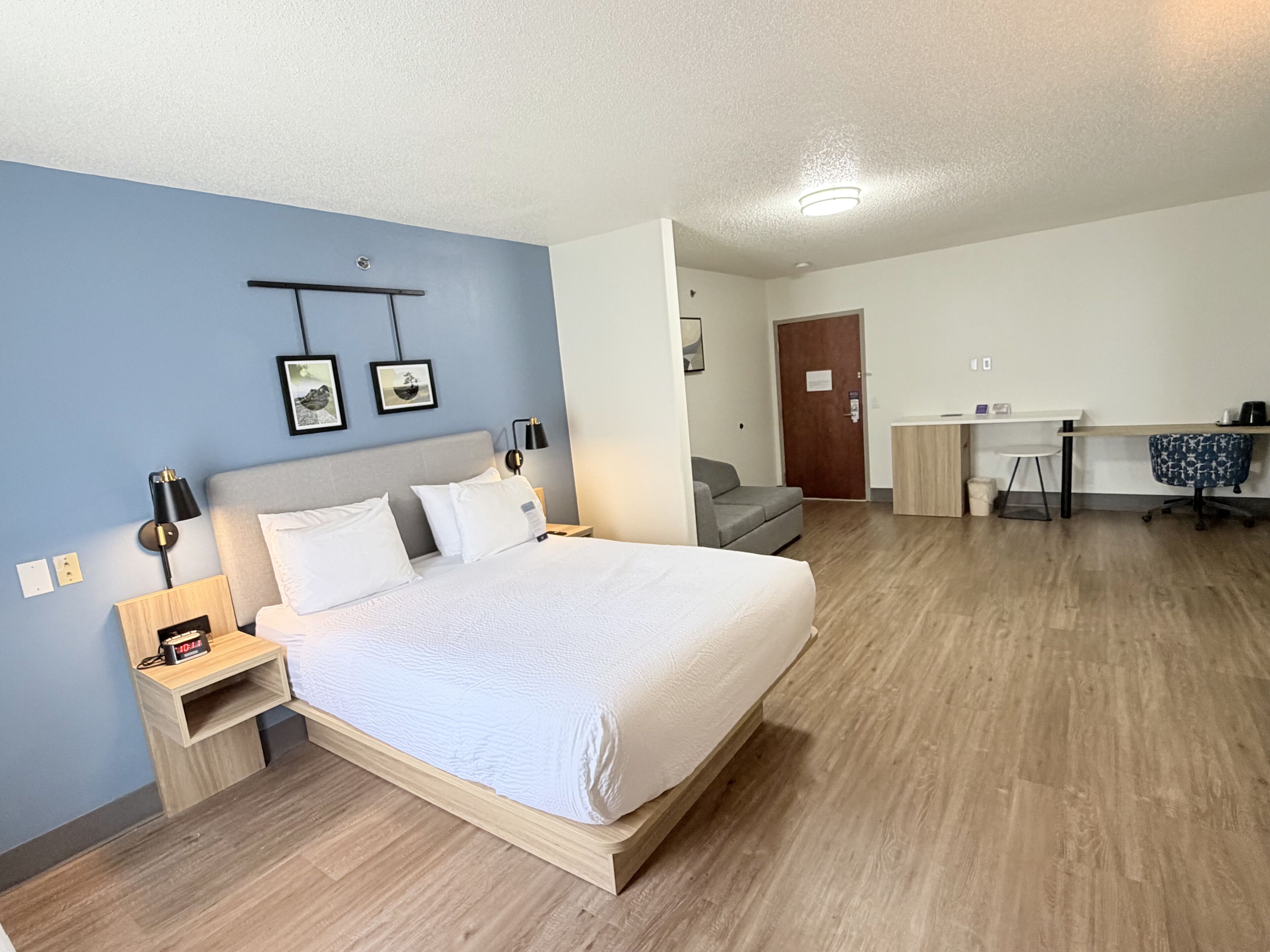 A hotel room with a bed, bedside table, lamp, wall art, a desk, chair, and a door.