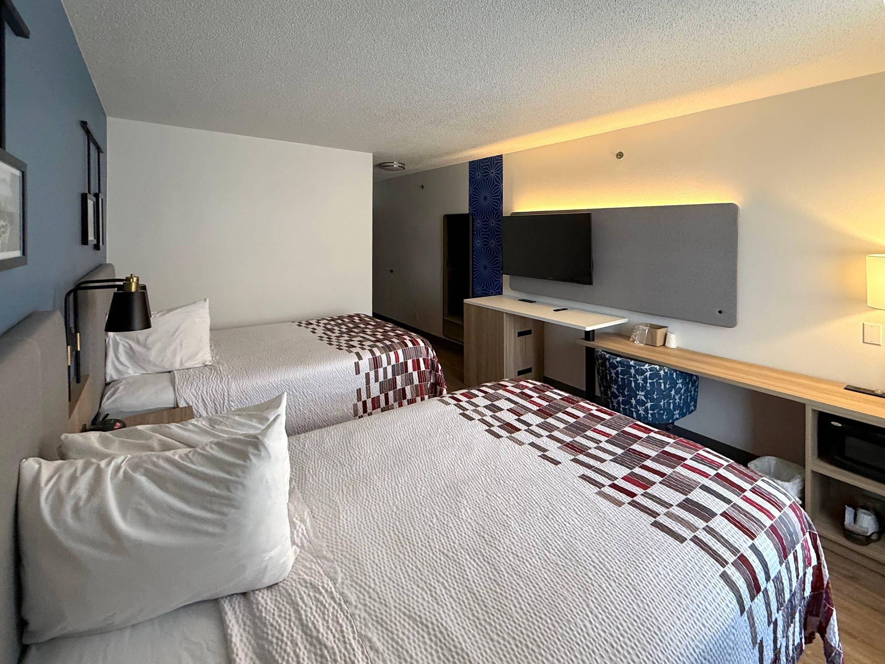 A hotel room with two beds, a desk, a TV, and a wall-mounted shelf.