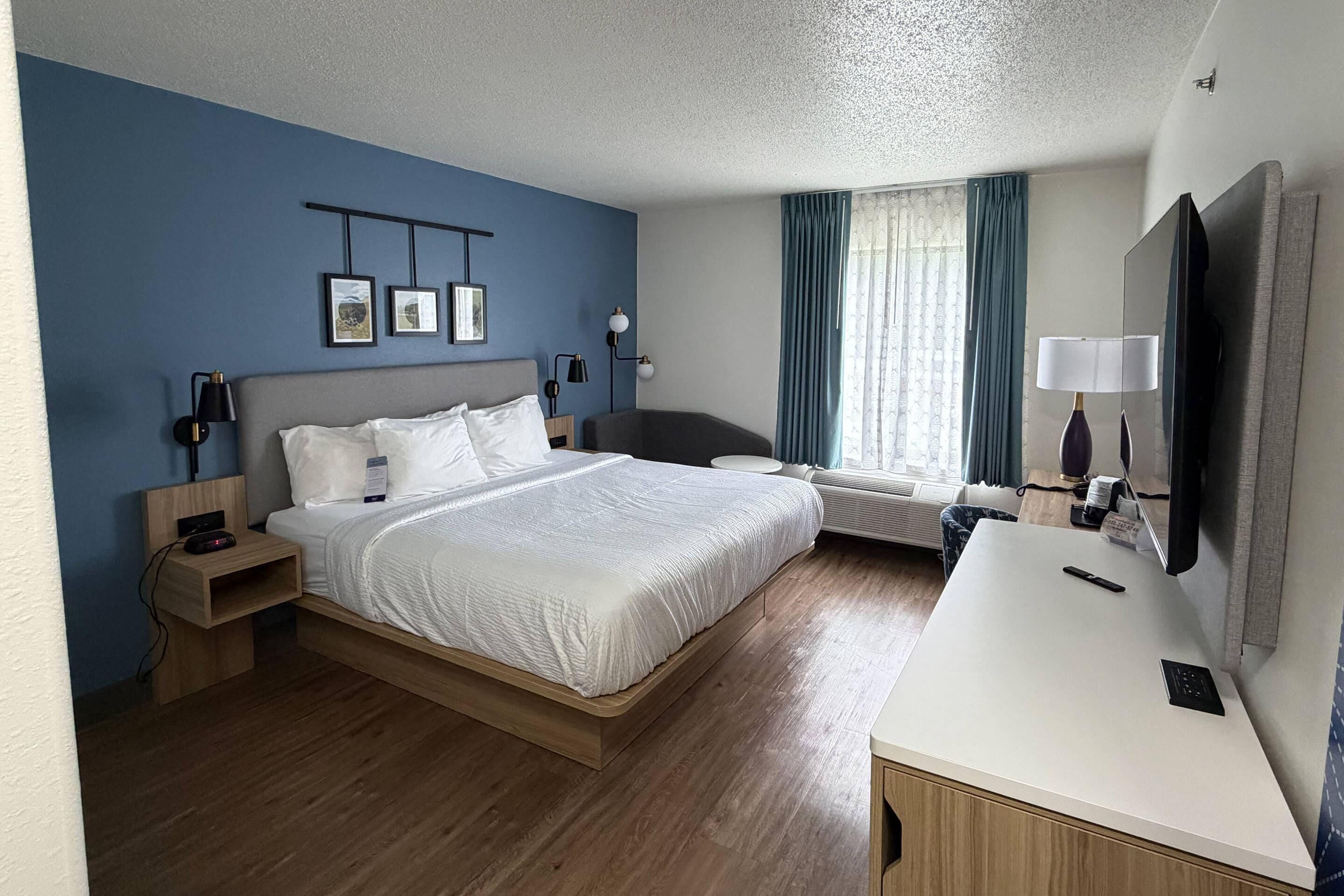 Una habitación de hotel con cama, mesitas de noche, escritorio, silla, televisor y ventana con cortinas.
