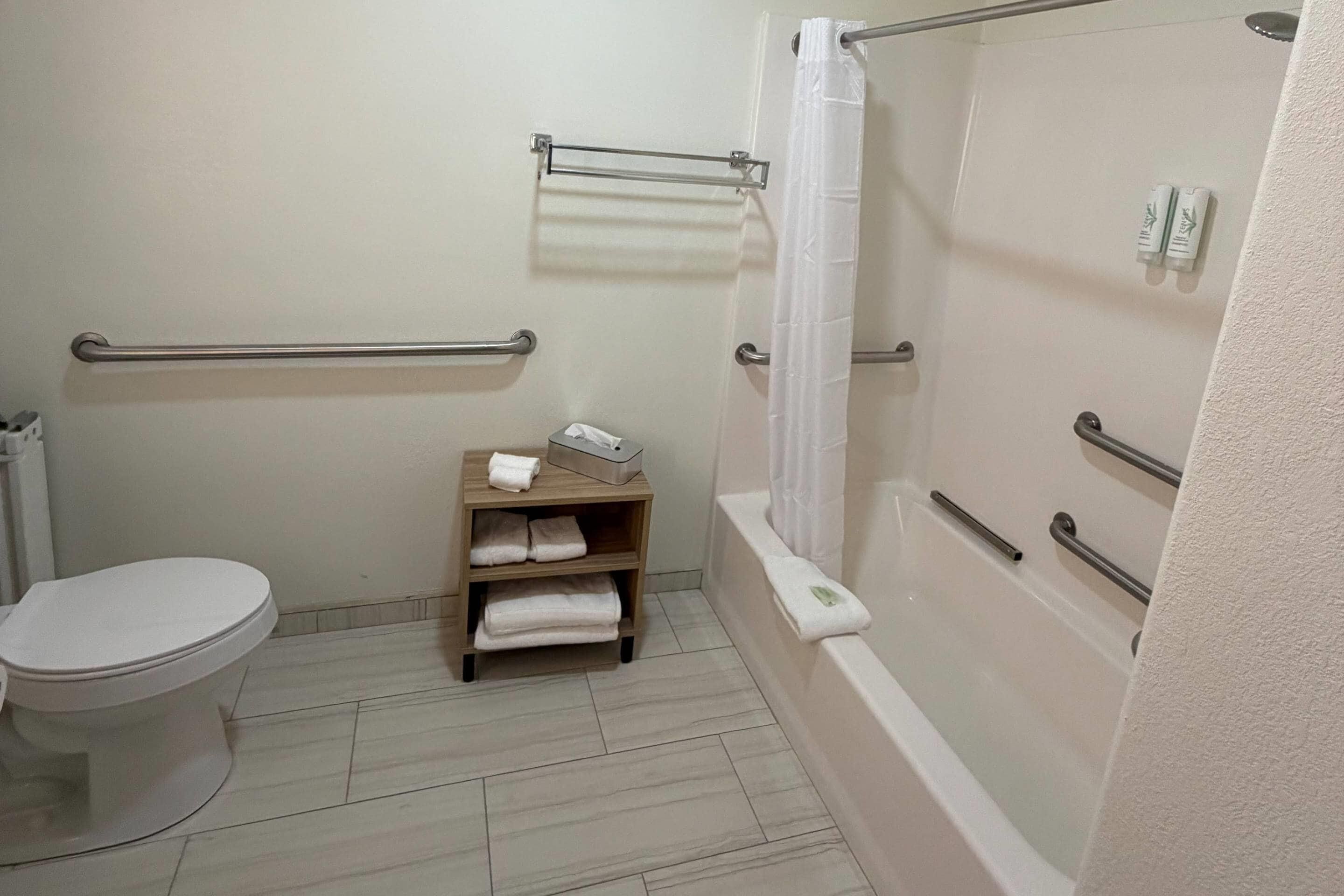 A bathroom with a toilet, a shelf with towels, a bathtub with a shower curtain, and grab bars.