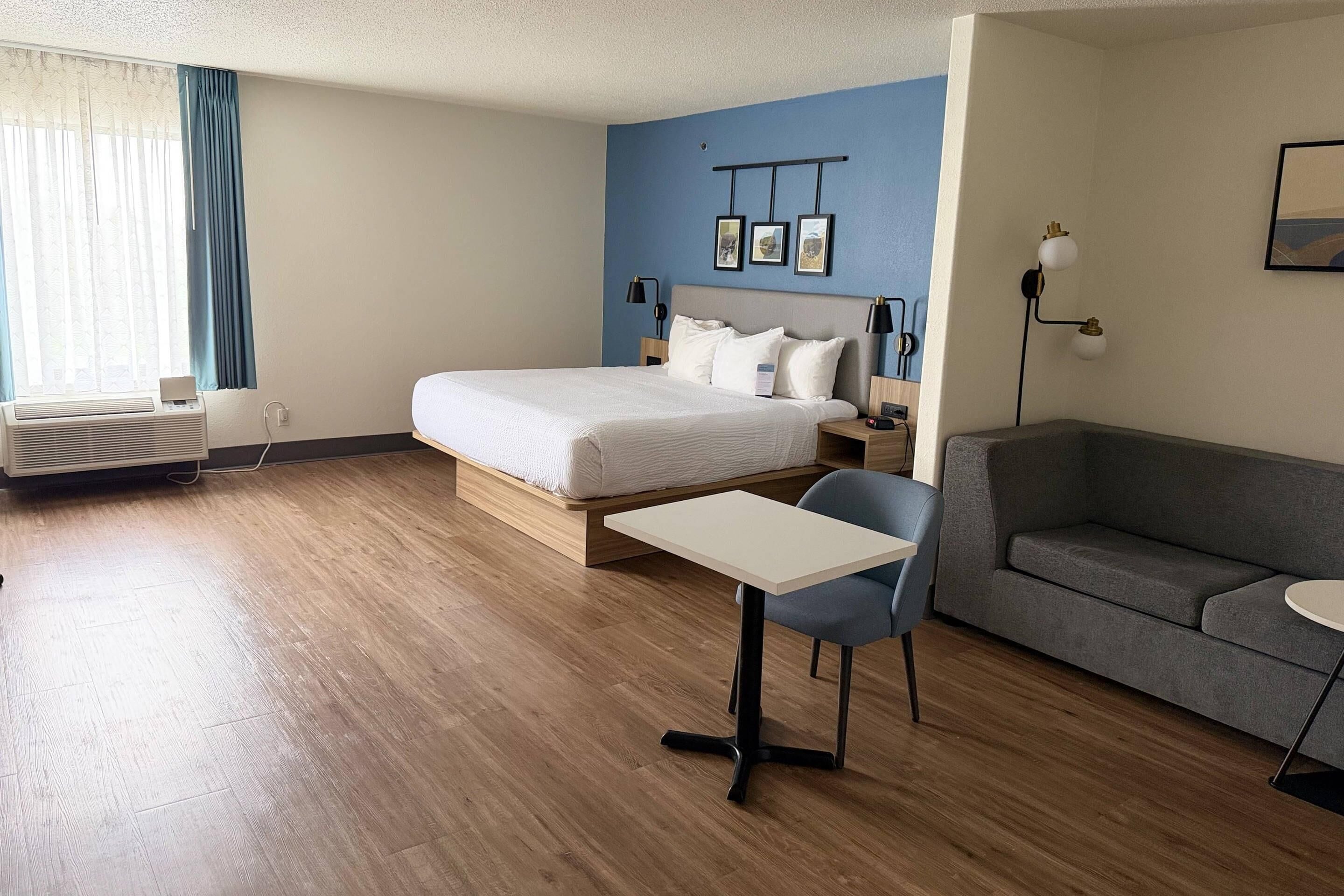 A hotel room with a bed, a desk, a chair, a sofa, a wall-mounted lamp, and a picture frame.