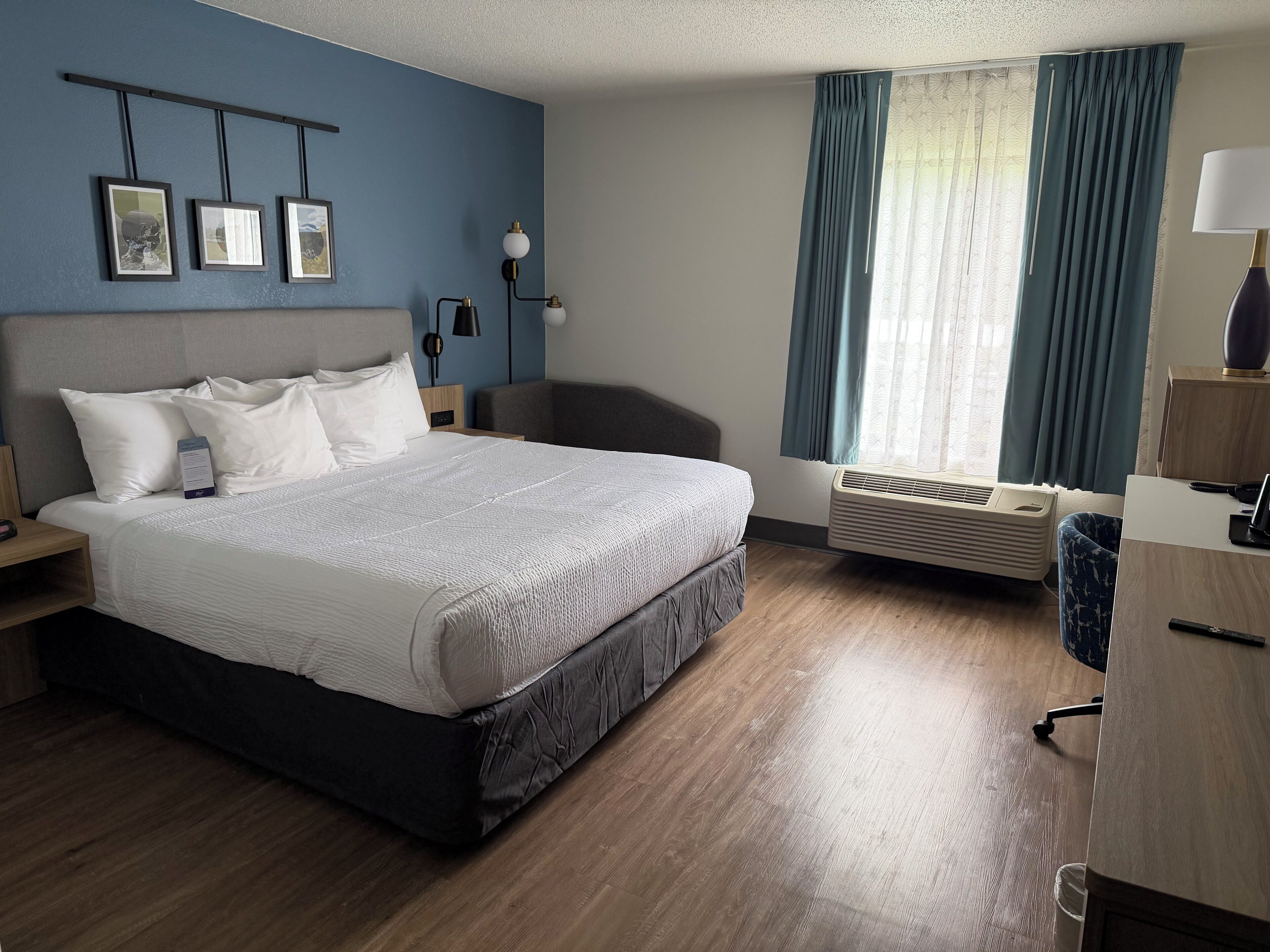 Une chambre d’hôtel comprenant un lit, un bureau, une chaise, une fenêtre avec des rideaux, un climatiseur fixé au mur et des tableaux encadrés accrochés au mur.
