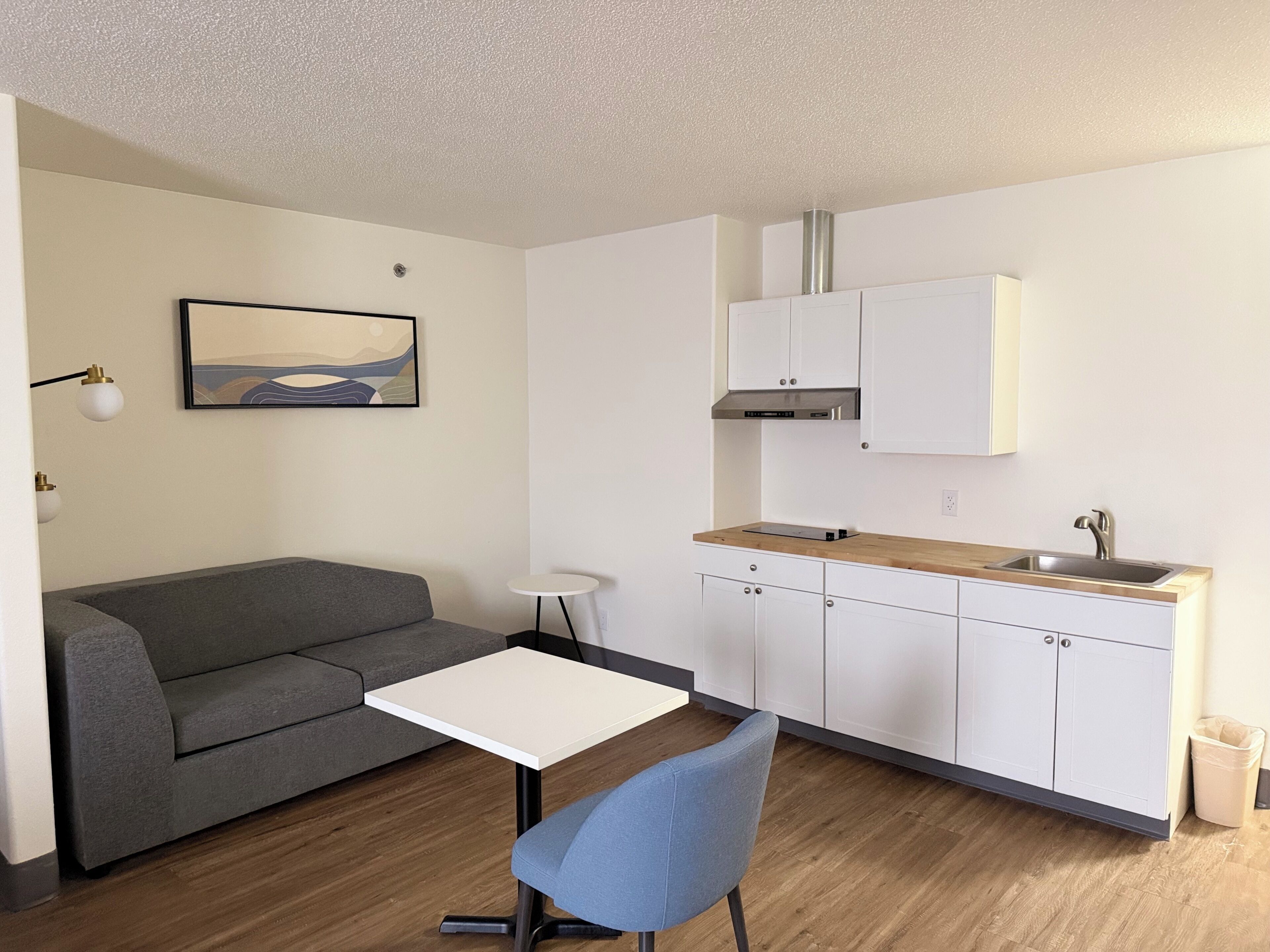 Um espaço compacto para viver, com cozinha compacta, sofá cinza, mesa pequena branca, cadeira azul e um quadro na parede.