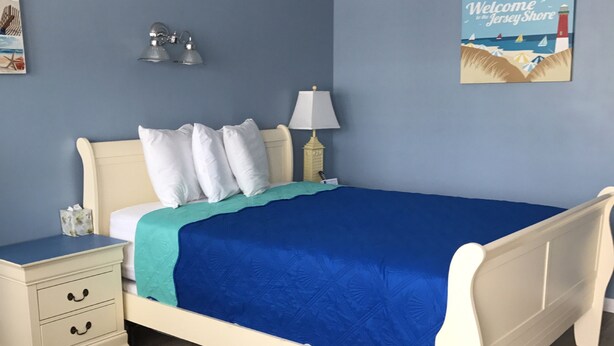 A bedroom with a bed, bedside tables, a nightstand, a lamp, and a wall decoration. - Pelican Point Motel (Point Pleasant Beach)