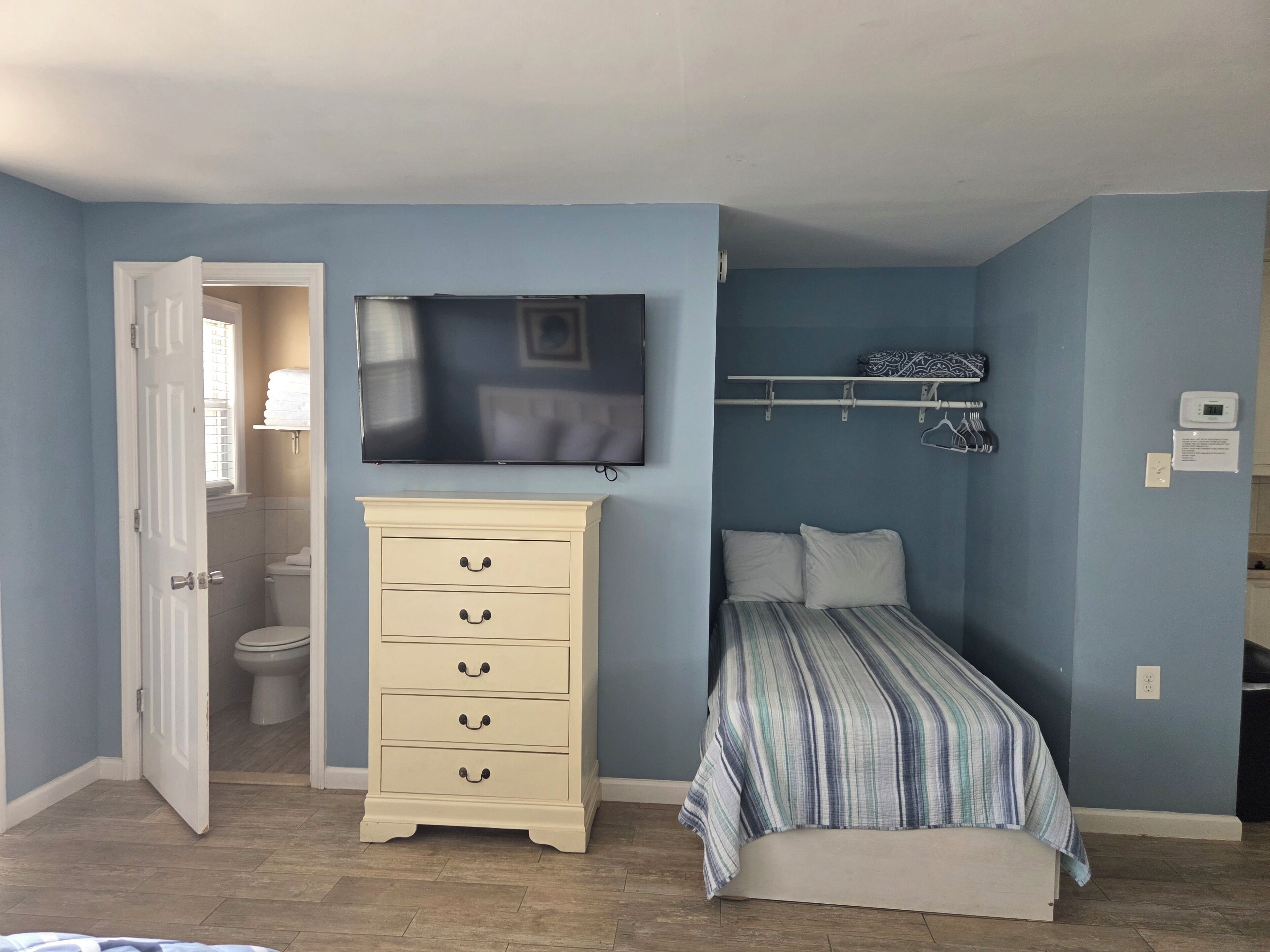 Une chambre à coucher comprenant un lit, une commode, un téléviseur fixé au mur et une porte donnant accès à une salle de bain.