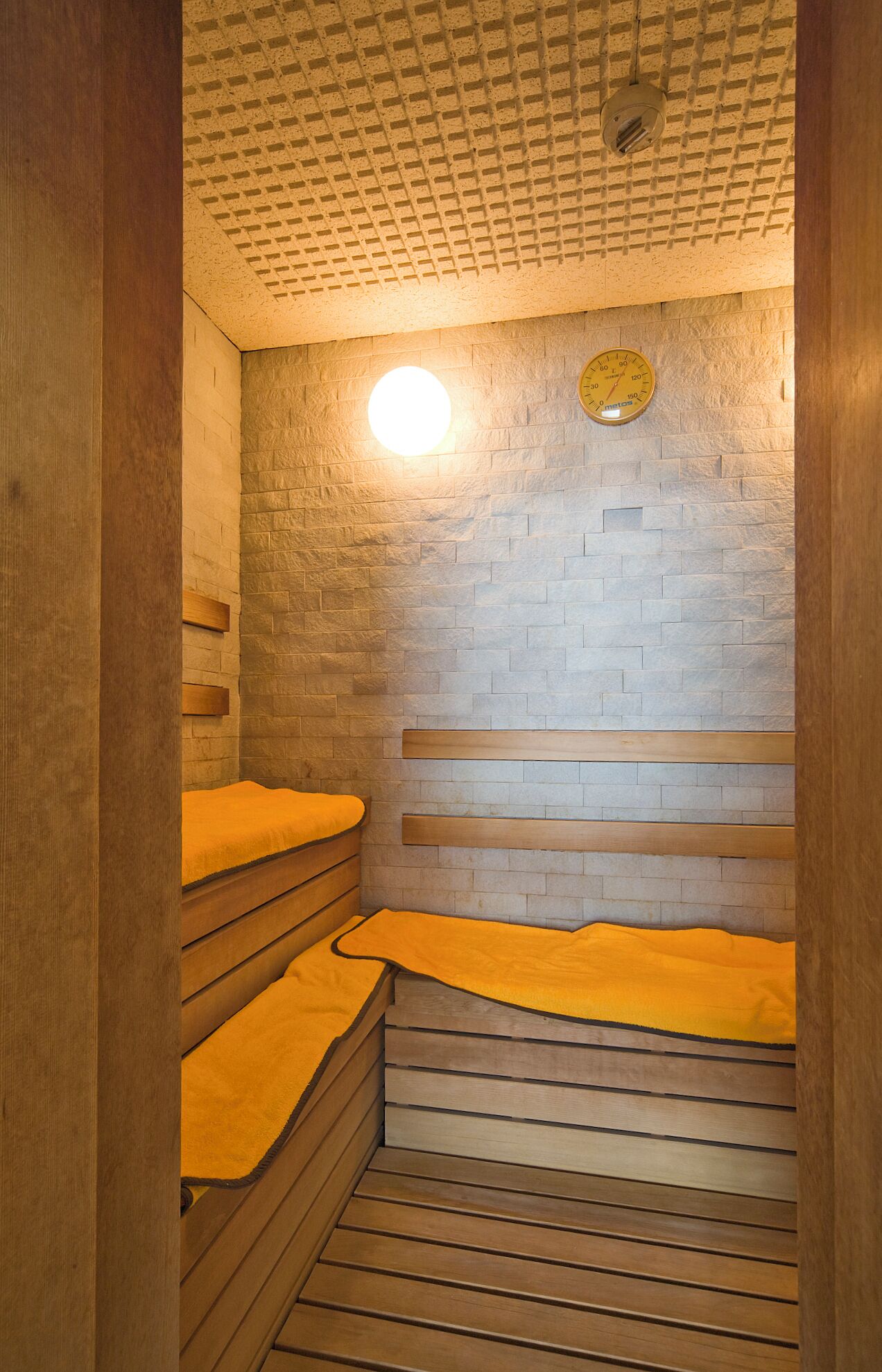 Eine Sauna mit Holzbänken und einer Ziegelwand, ausgestattet mit einer runden Leuchte und einem an der Wand befestigten Thermometer.
