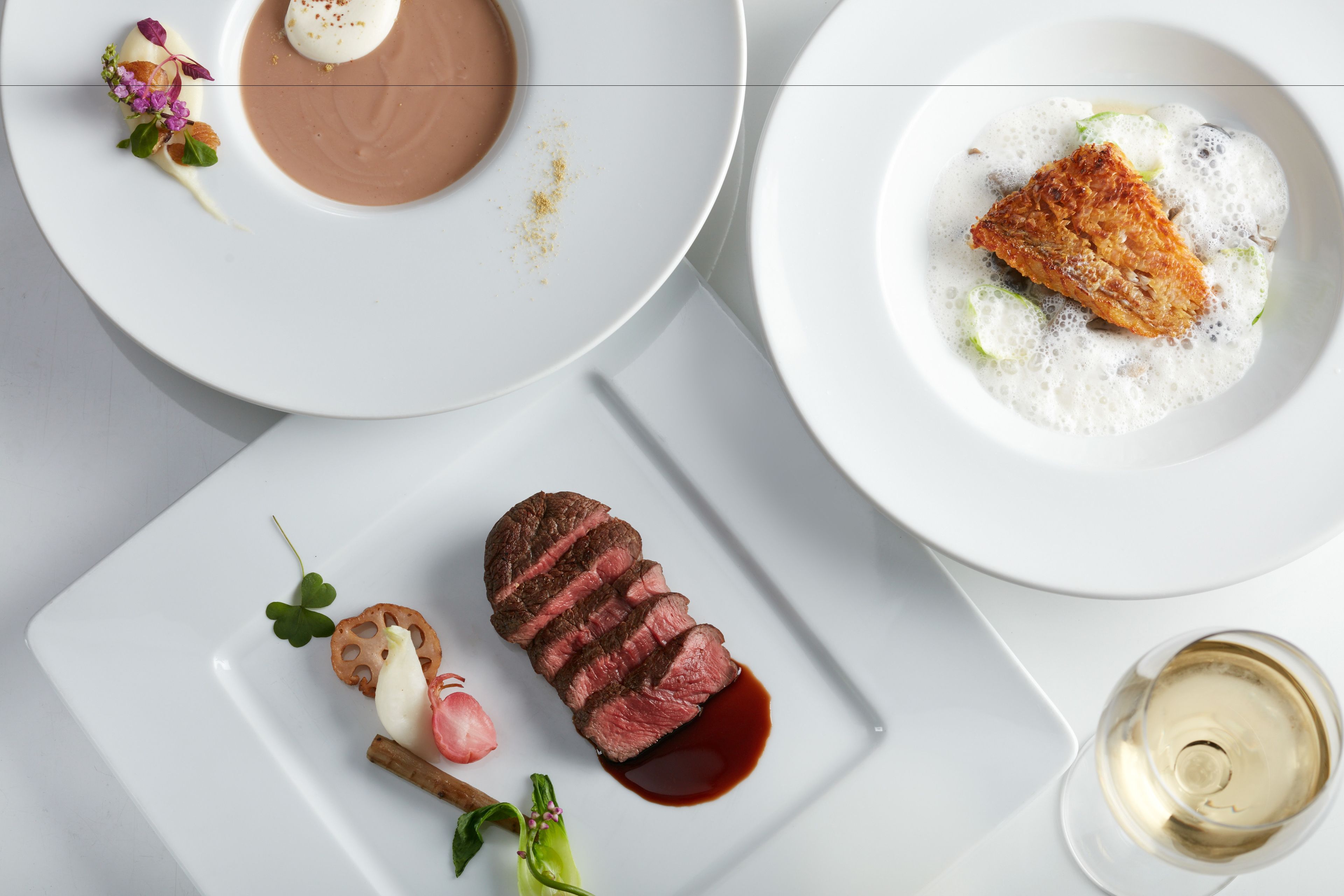 Une assiette blanche avec un morceau de viande, un petit récipient contenant une sauce et un verre de vin blanc.