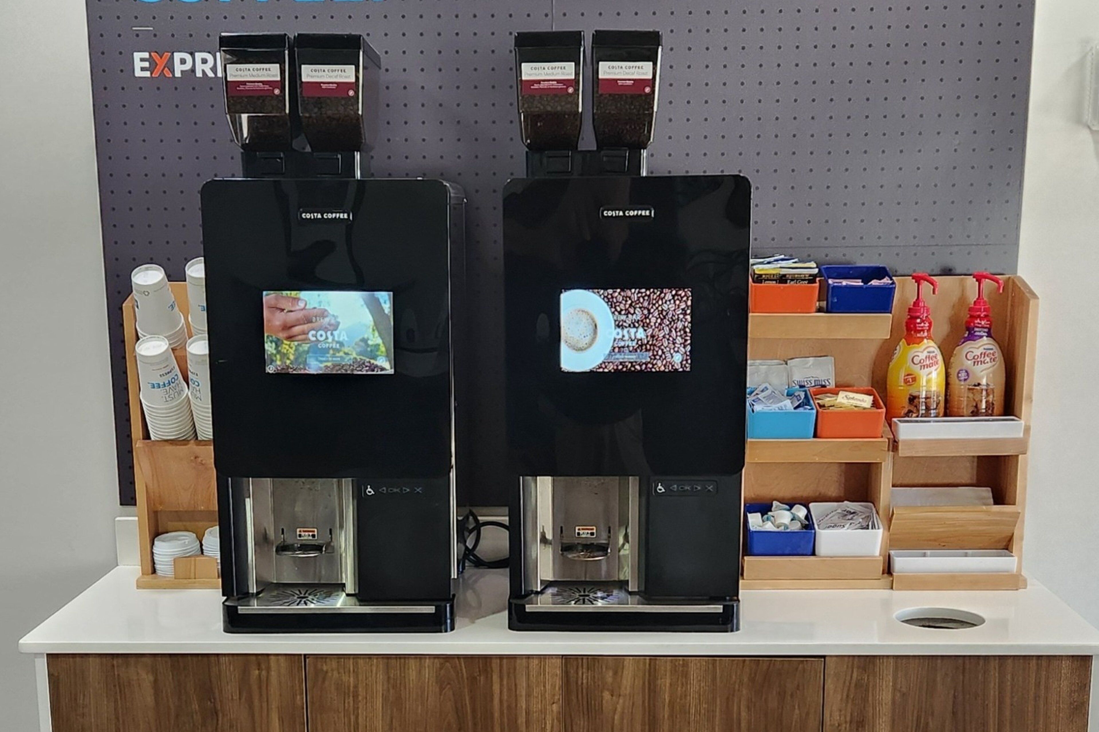 Two coffee machines with digital displays, a shelf with disposable cups, and a wall-mounted display with the word 'EXPRESS'.