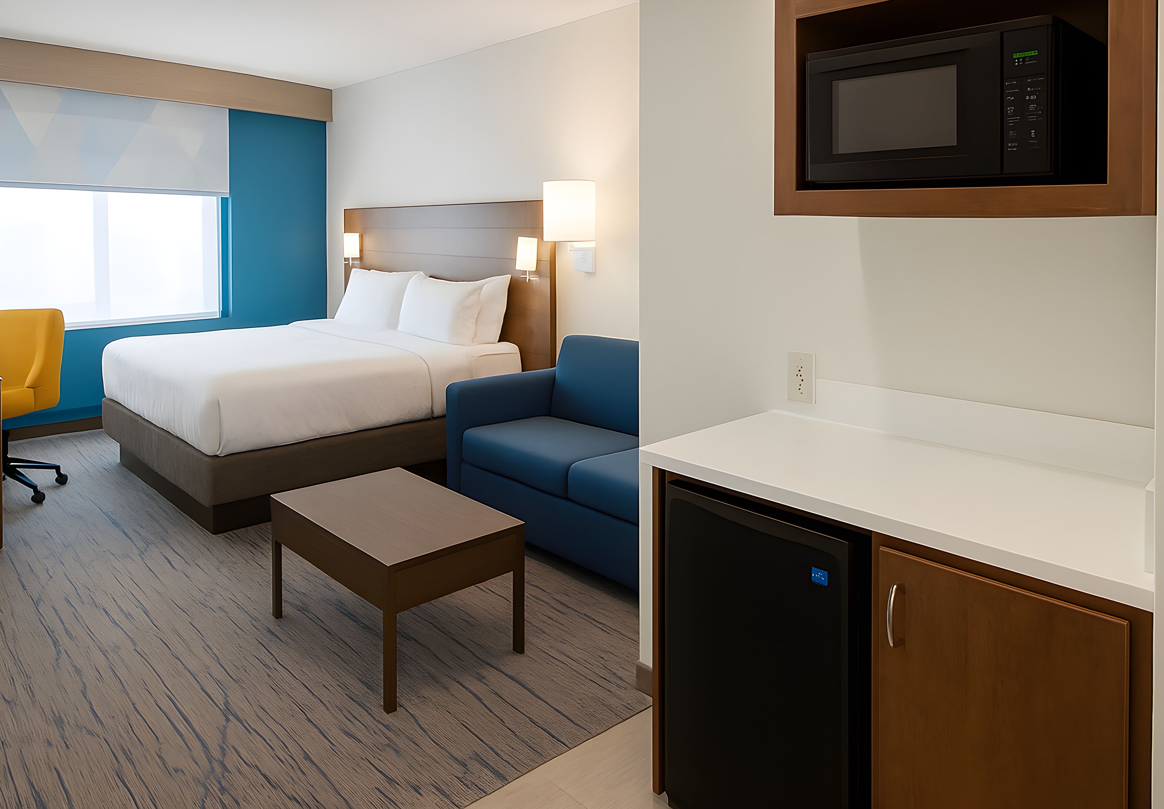 A hotel room with a bed, a desk, a chair, a microwave, and a refrigerator.