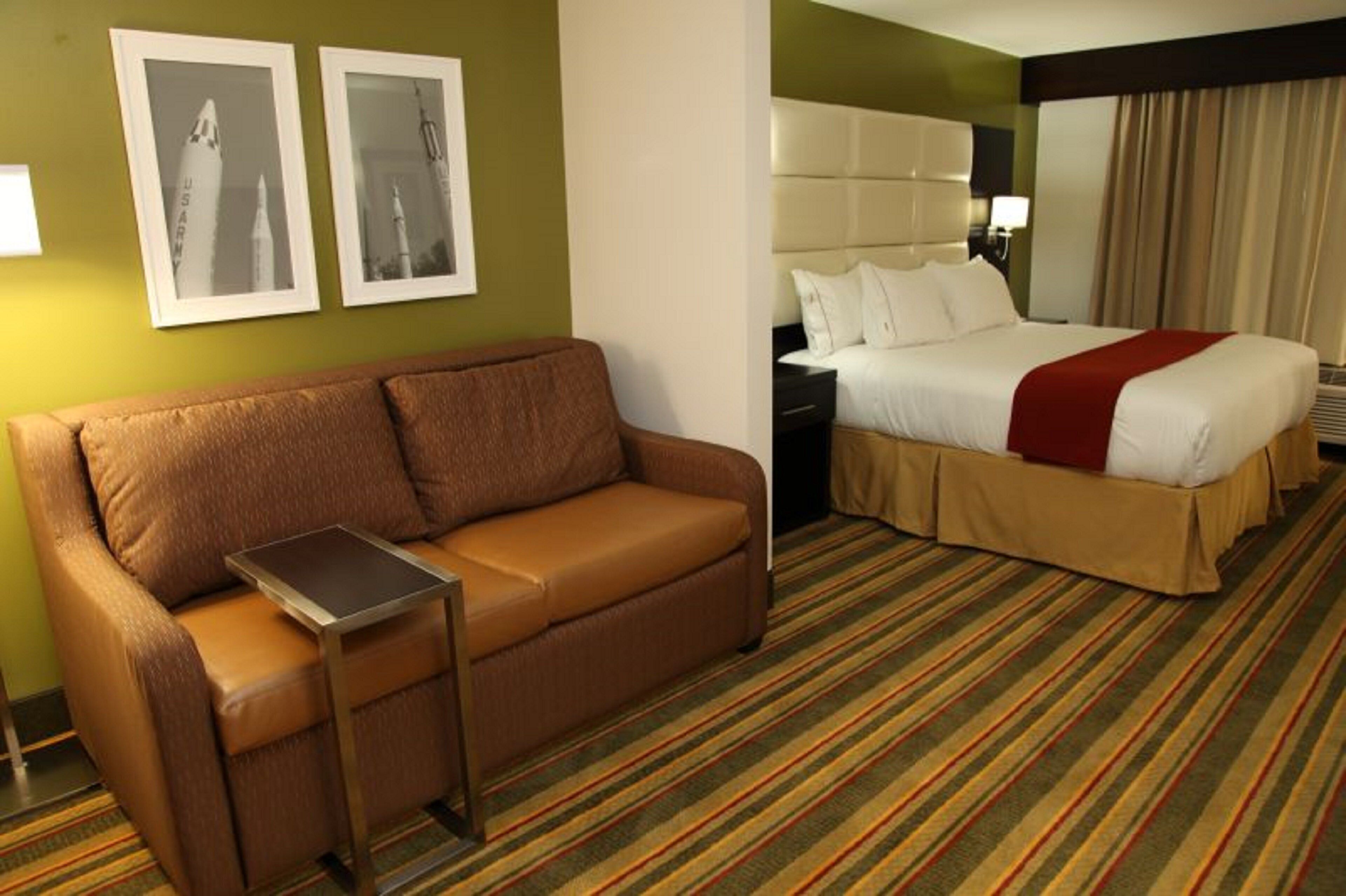 A hotel room with a brown sofa, a bed with white bedding and a red throw, and two framed pictures on the wall.