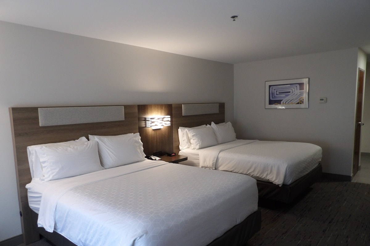 Habitación de hotel con dos camas, una mesita de noche, una lámpara de pared y un cuadro enmarcado.