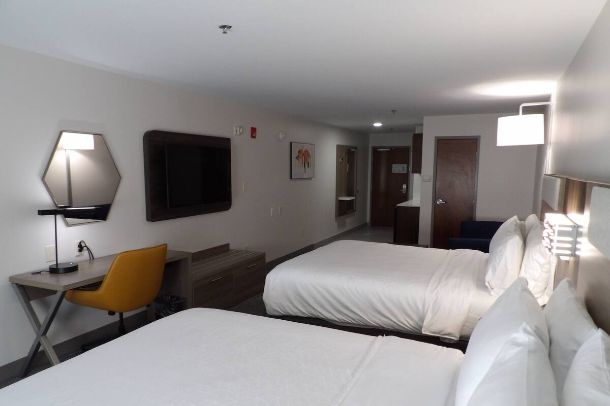 Habitación de hotel con dos camas, un escritorio con silla, una lámpara y un televisor en la pared.