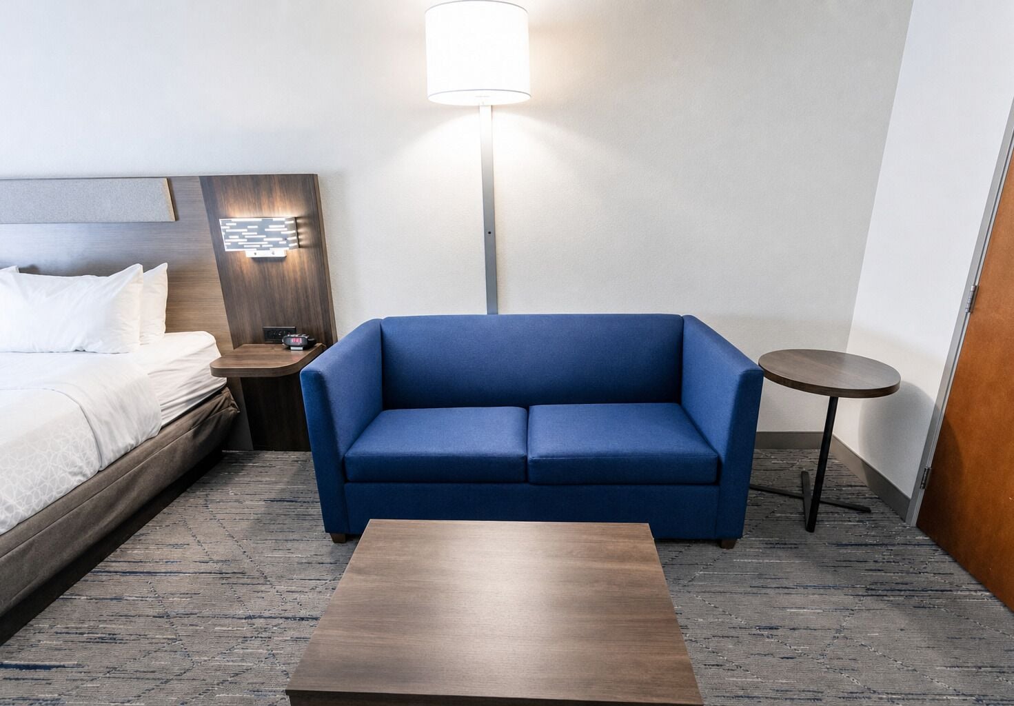 A hotel room with a bed, a blue sofa, a small table, a nightstand, and a floor lamp.