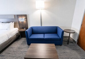 A hotel room with a bed, a blue sofa, a small table, a nightstand, and a floor lamp. - Holiday Inn Express & Suites Huntsville Airport by IHG (Madison)