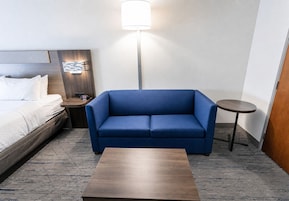 Quarto de hotel com uma cama, um sofá azul, uma mesinha, um criado-mudo e um abajur.