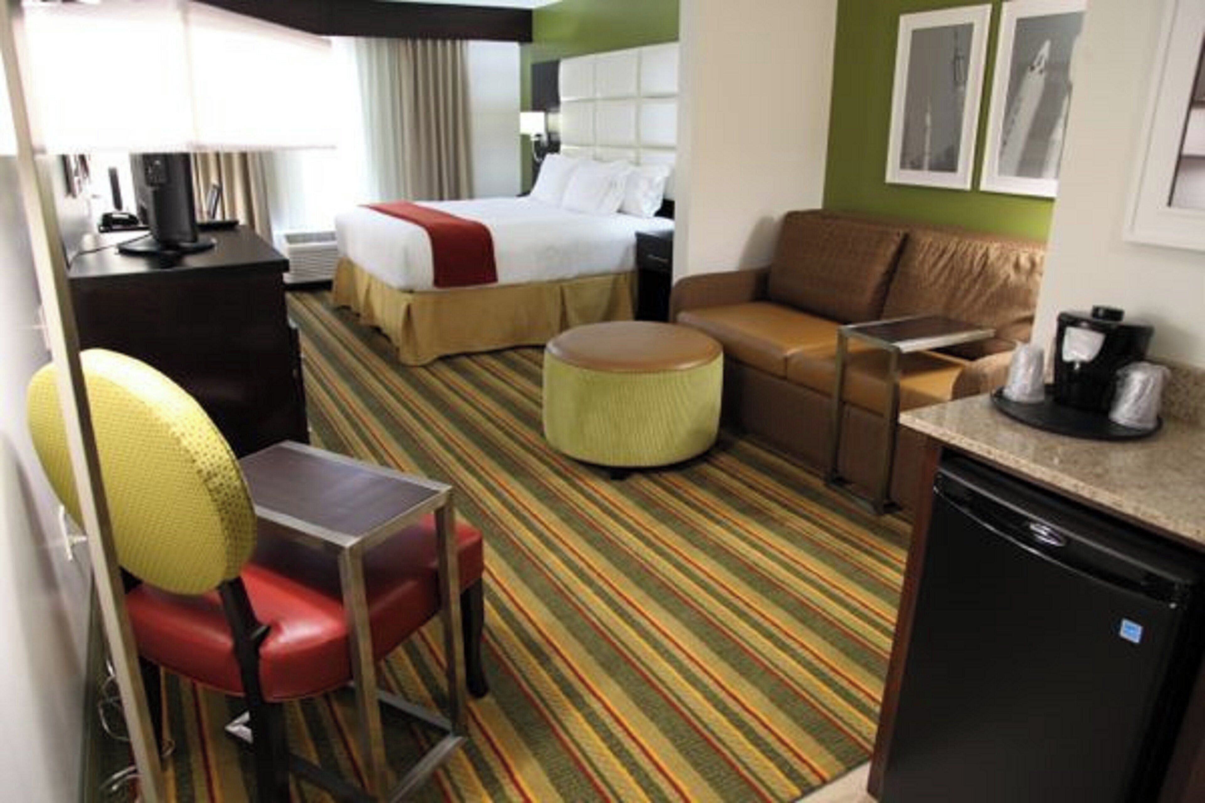 A hotel room with a bed, a sofa, a desk with a chair, a coffee maker, and a small kitchenette.