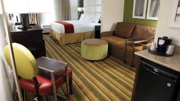 A hotel room with a bed, a sofa, a desk with a chair, a coffee maker, and a small kitchenette.