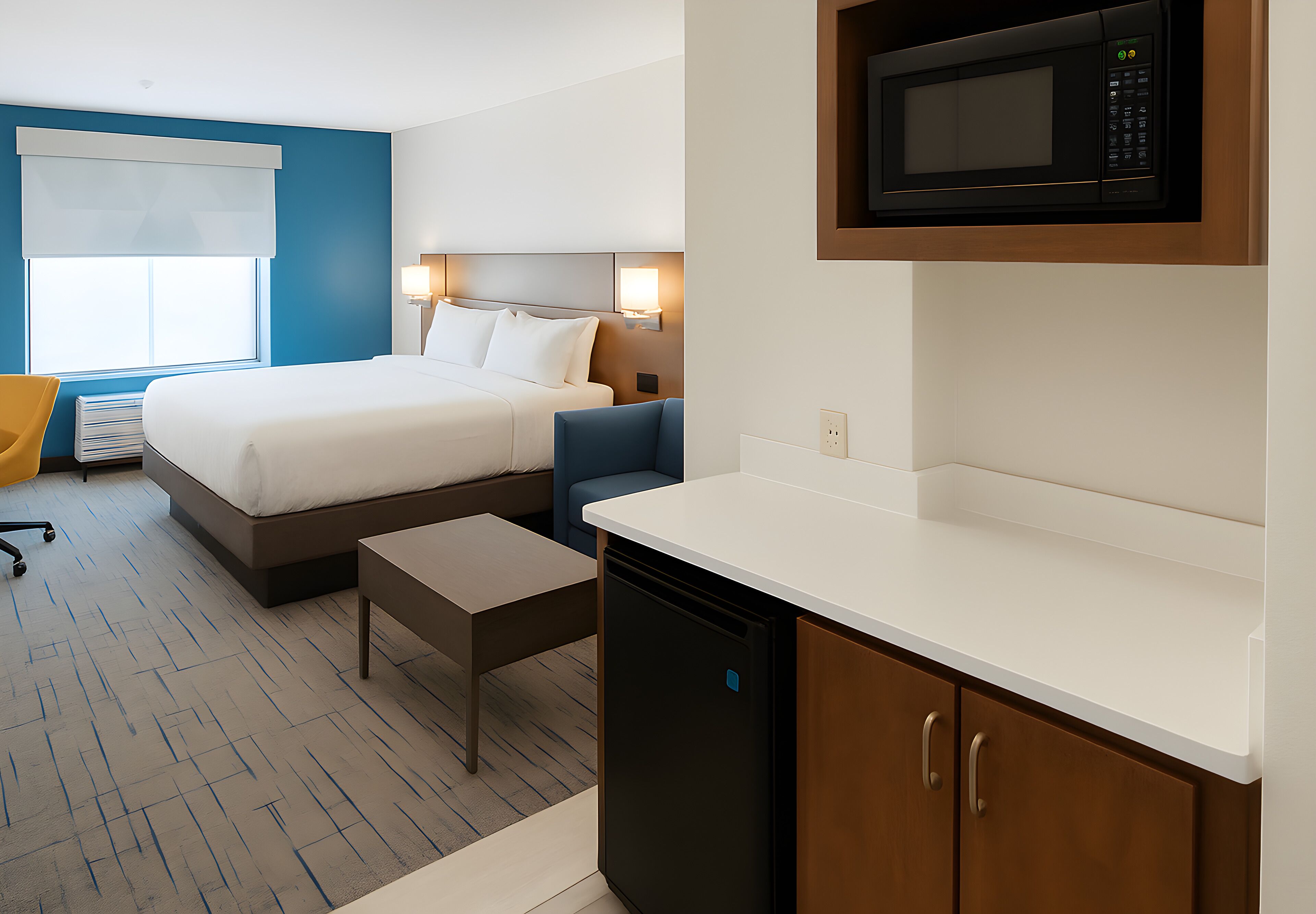 A hotel room with a bed, bedside table, chair, and a kitchenette area with a microwave.