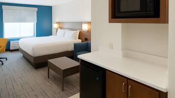 A hotel room with a bed, bedside table, chair, and a kitchenette area with a microwave.