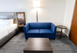 A hotel room with a bed, a blue sofa, a small table, a lamp, and a wall-mounted clock. - Holiday Inn Express & Suites Huntsville Airport by IHG (Madison)