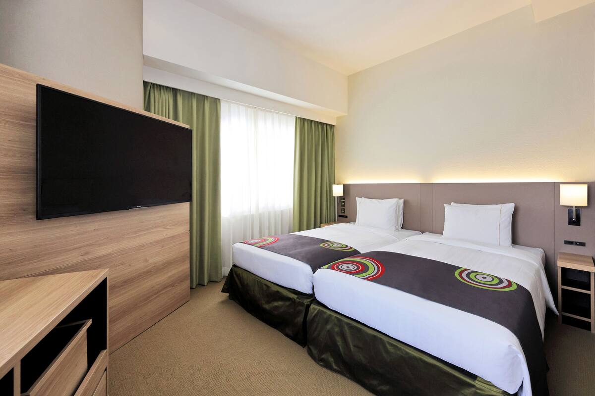 Ein Hotelzimmer mit zwei Betten, einem an der Wand befestigten Fernseher, einem Schreibtisch und einem Nachttisch mit Lampe.