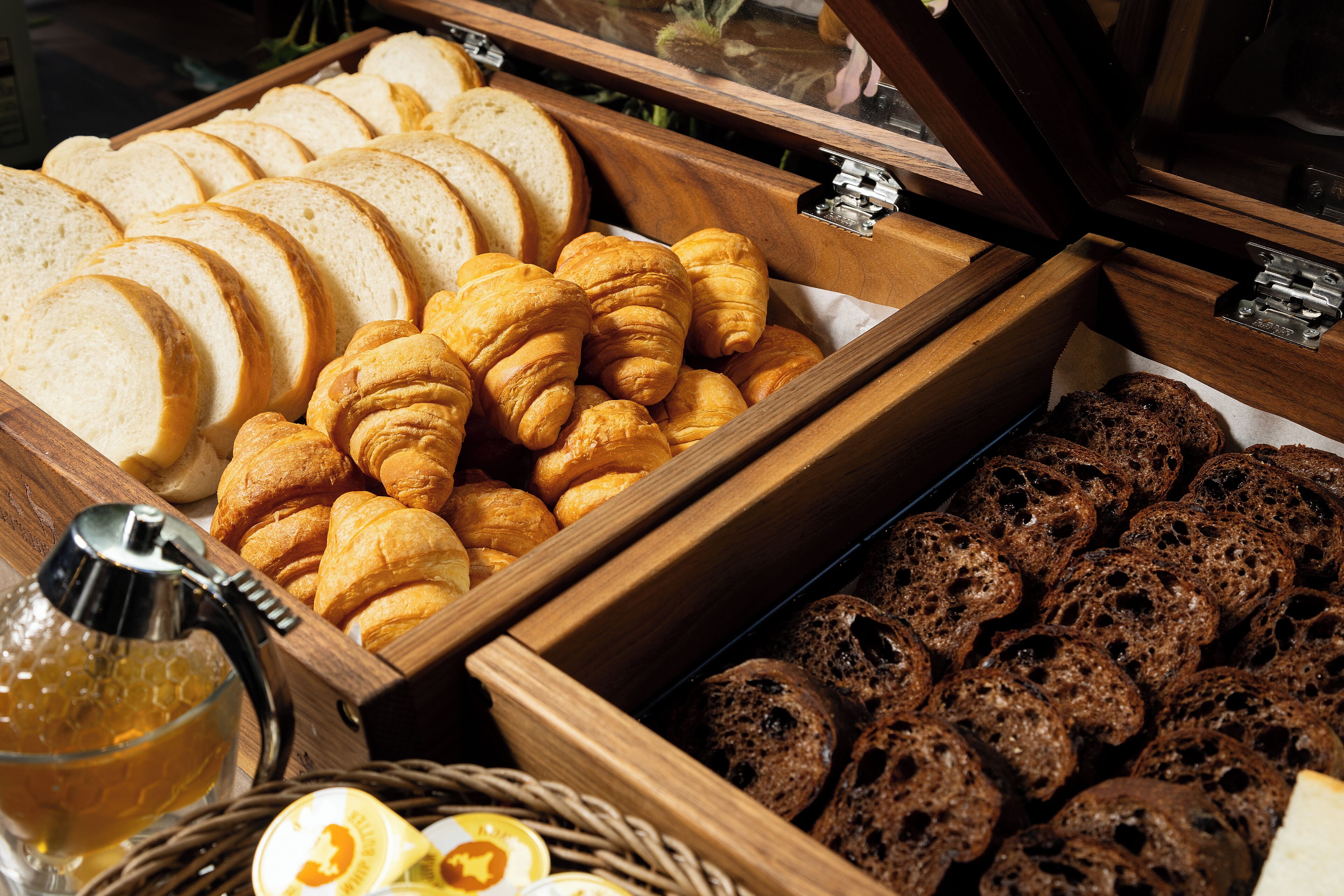A wooden buffet tray with sliced bread, croissants, and dark bread rolls.